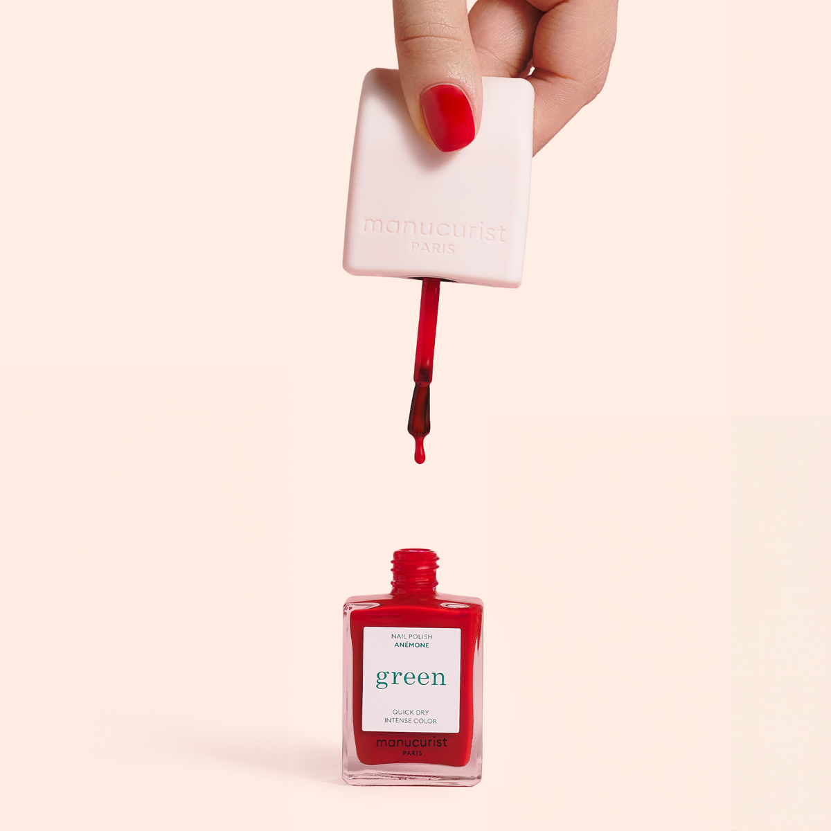 A hand with red-painted nails holds a brush applicator above an open bottle of red nail polish labeled green by Manucurist Paris, against a pale background.
