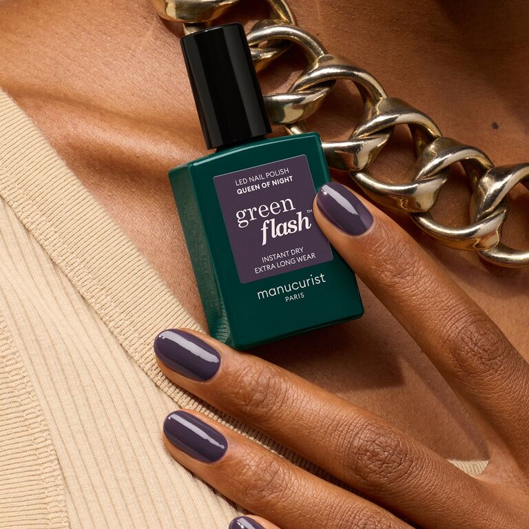 A hand with dark purple-gray nails holds a bottle of Manucurist Queen of Night polish, wearing a chunky gold chain necklace against a soft beige fabric background.
