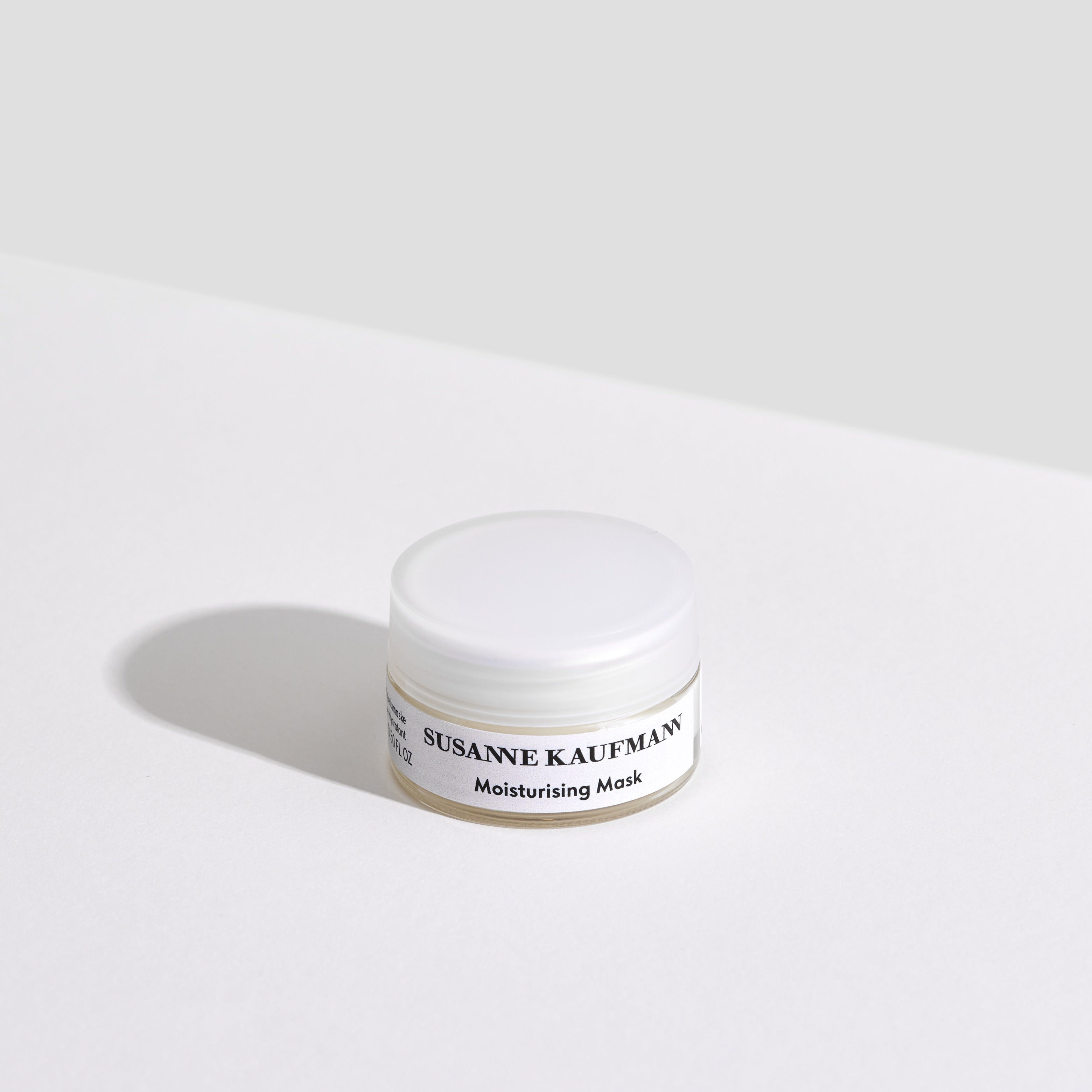 A small, white jar of Susanne Kaufmann Moisturising Mask sits on a white surface, casting a soft shadow. The background is light grey and minimalistic, highlighting the product.