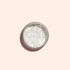 The Manucurist Pearlescent Glitter Flakes come in a small round container on a pale peach background, casting a soft shadow—ideal for adding dazzling shimmer to your next manicure.