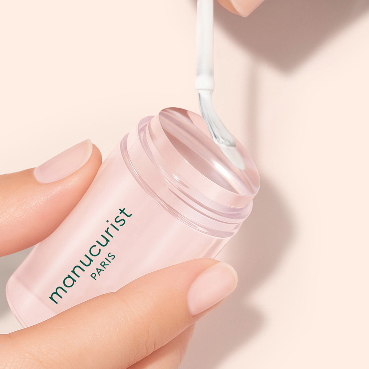 A hand with light nude nails holds a pink container labeled manucurist PARIS while another hand uses a brush to collect product from the open container.
