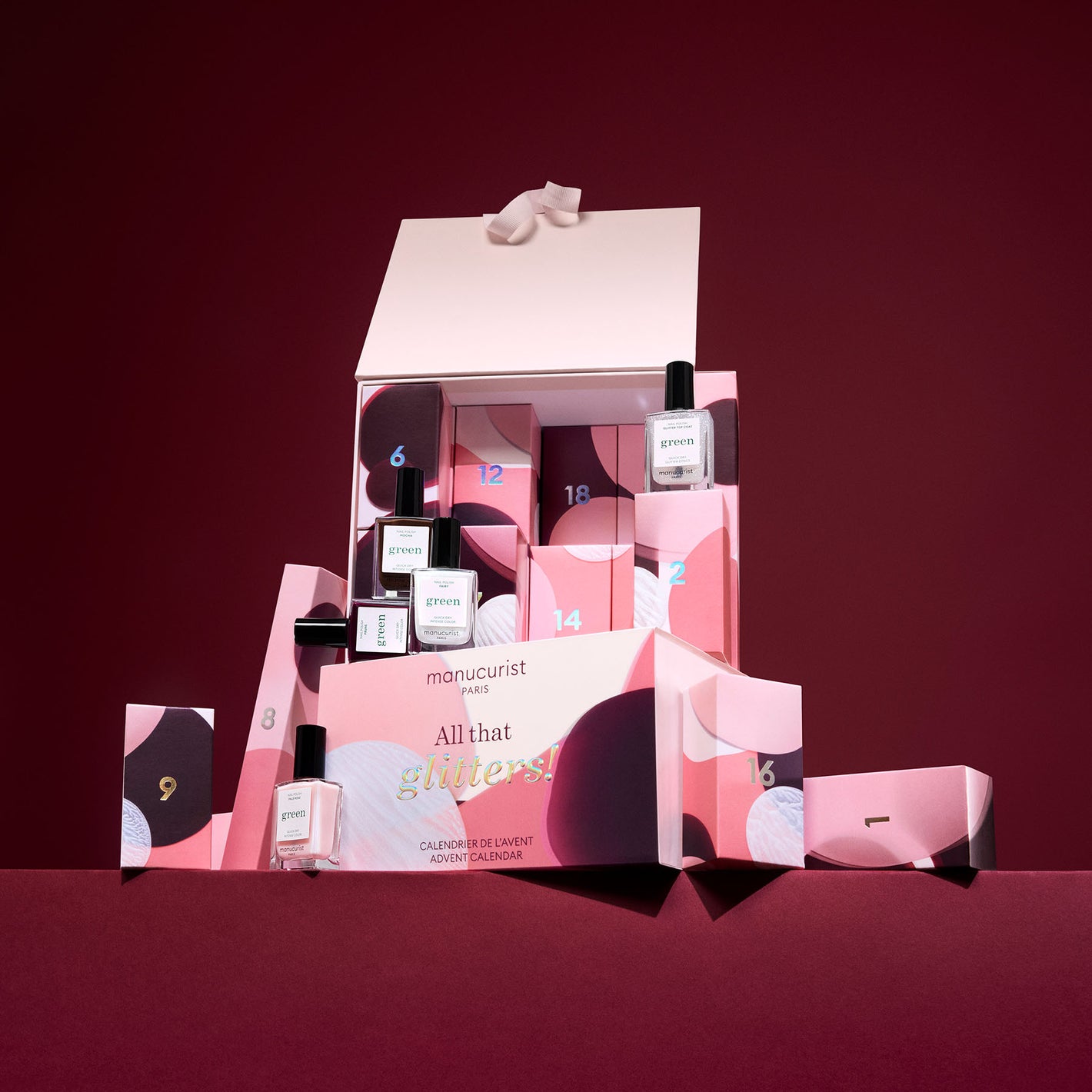 A pink and red advent calendar box with numbered compartments, some open to reveal nail polish bottles, sits on a red surface against a deep red background. A white lid with a bow is propped open on top.