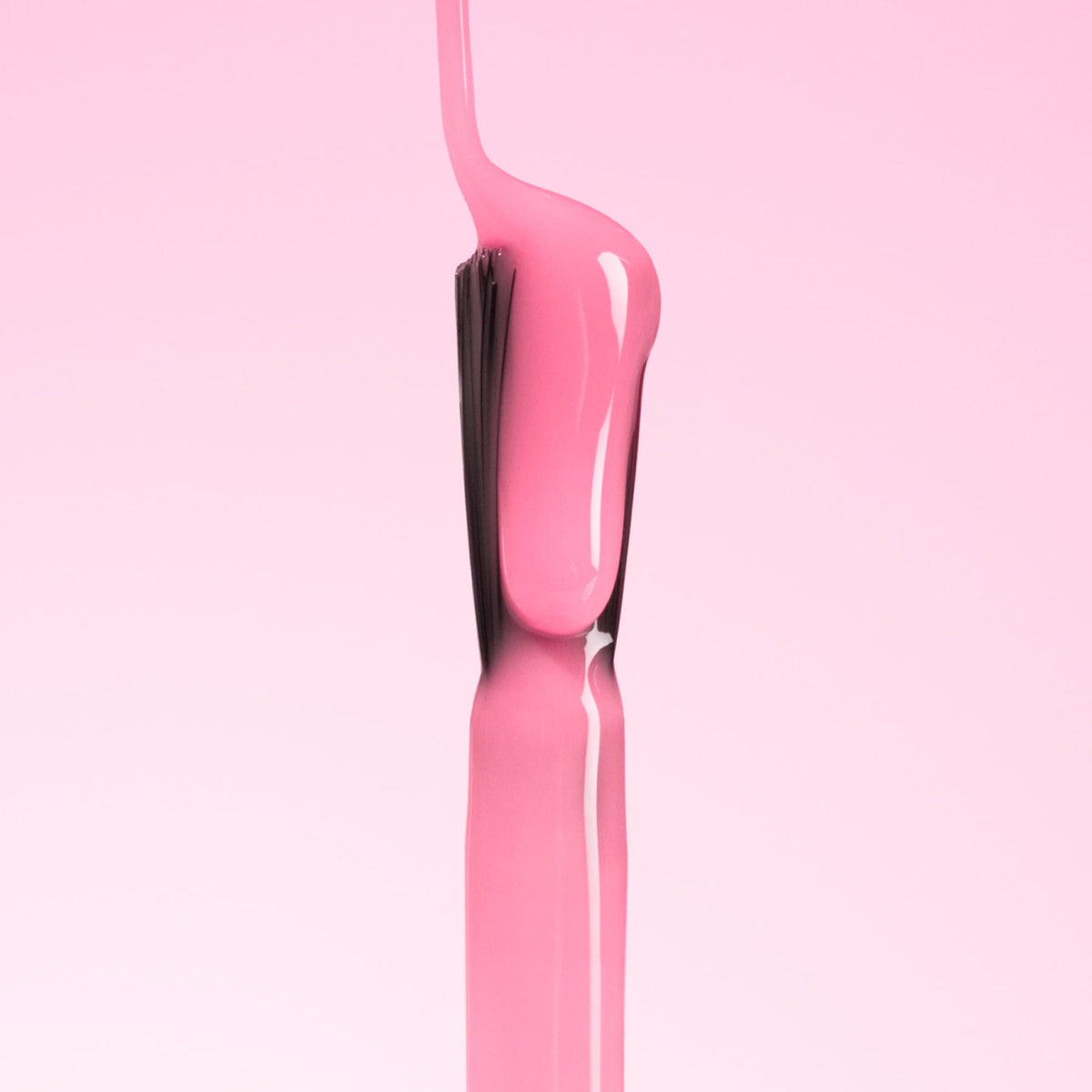 A close-up of a brush being pulled out of a bottle, coated in glossy pink nail polish against a solid pink background.