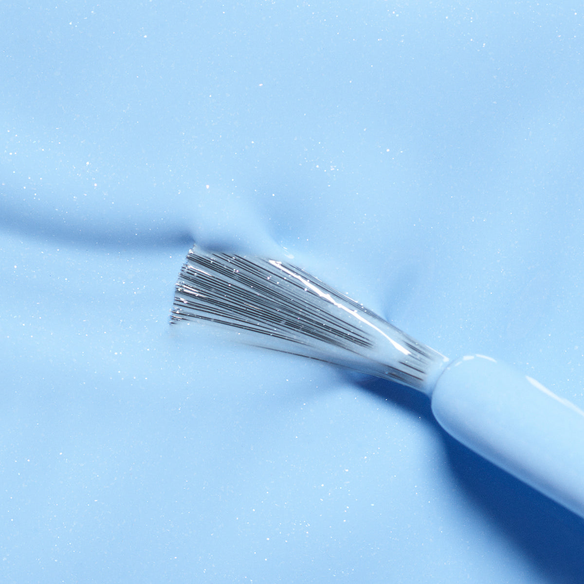 A close-up of a brush with silver bristles applying Manucurist’s Pomme the Fish, a light blue, non-toxic nail polish, on a smooth, glossy surface.