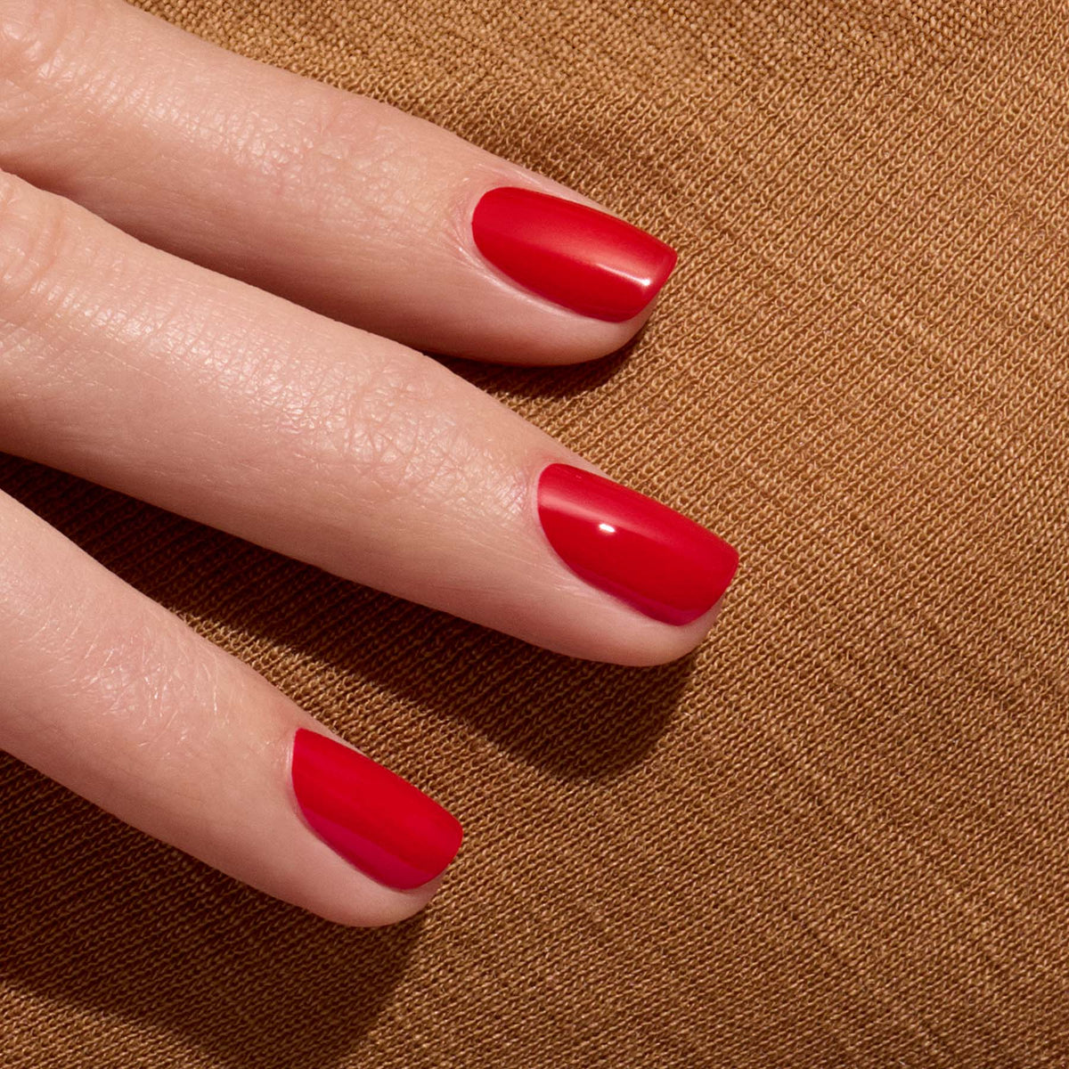 Gros plan sur une main aux ongles soigneusement manucurés, vernis avec le vernis Poppy Red brillant de Manucurist, posée sur un tissu brun texturé.