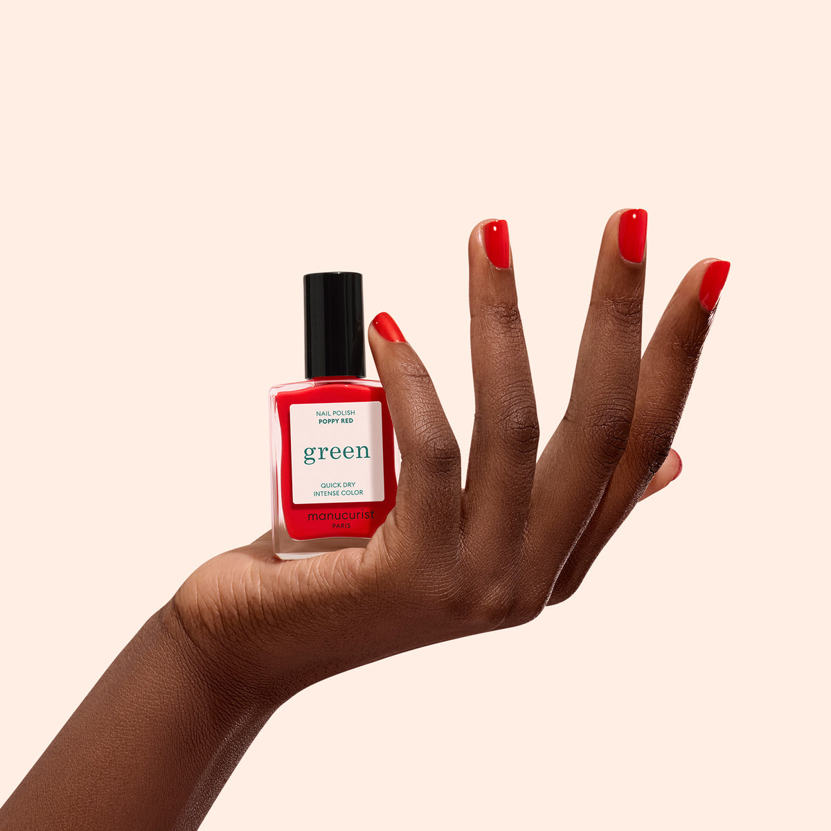 Une main aux ongles soigneusement vernis en Poppy Red tient un flacon de vernis à ongles Manucurist Poppy Red sur un fond beige clair uni.