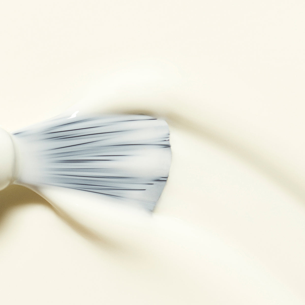 A close-up of a makeup brush spreading creamy white Porcelaine foundation from Manucurist on a smooth, light surface. The bristles are splayed out, evenly distributing the product.