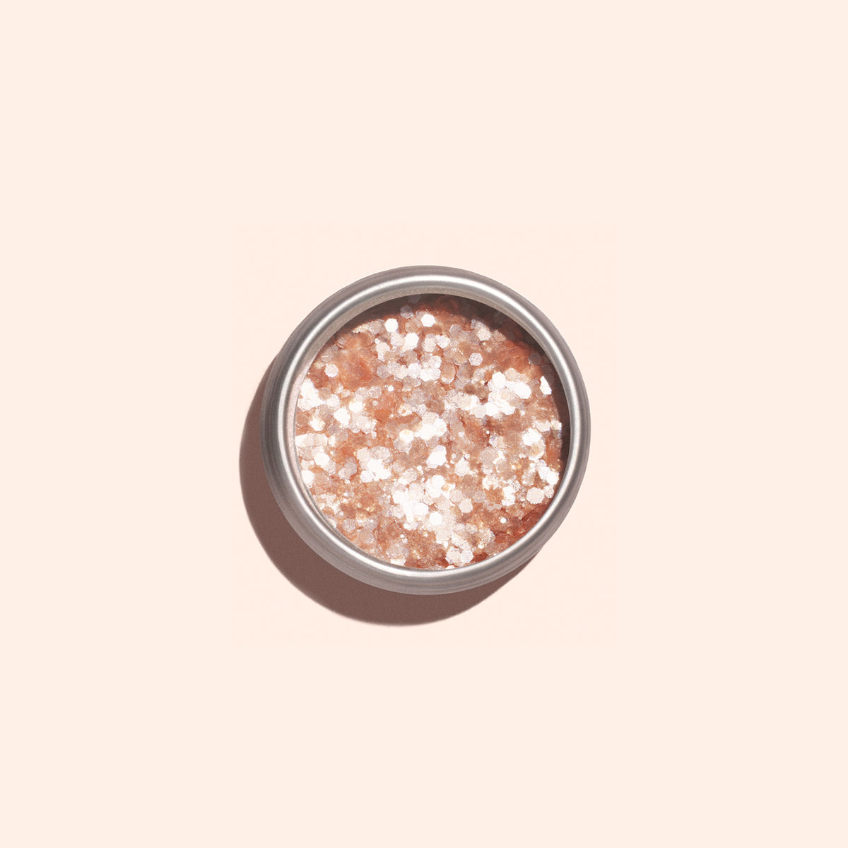 Une boîte ronde en métal de paillettes biodégradables Blush de Manucurist, contenant de gros sels de bain roses et blancs aux reflets rose pâle, est posée sur une surface beige clair et projette une ombre douce vers la gauche.
