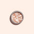 Une boîte ronde en métal de paillettes biodégradables Blush de Manucurist, contenant de gros sels de bain roses et blancs aux reflets rose pâle, est posée sur une surface beige clair et projette une ombre douce vers la gauche.