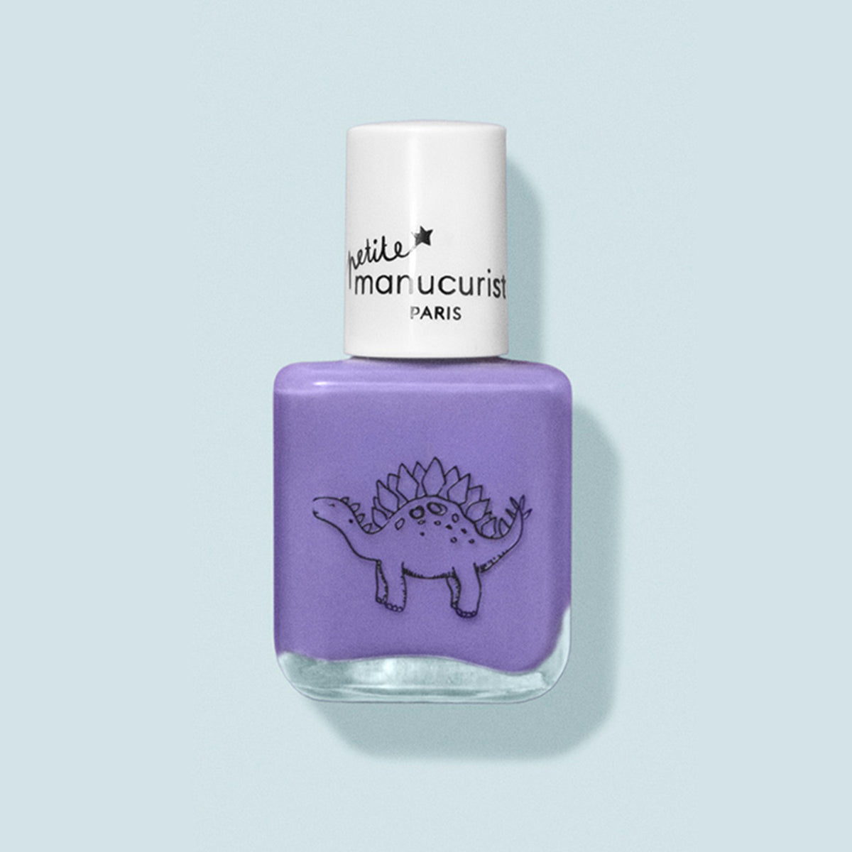 A bottle of purple kids nail polish named Hector the Stegosaurus by Manucurist with a white cap. It features a black line dinosaur drawing and “petite Manucurist PARIS” label. Made from natural ingredients. Background is pale blue.