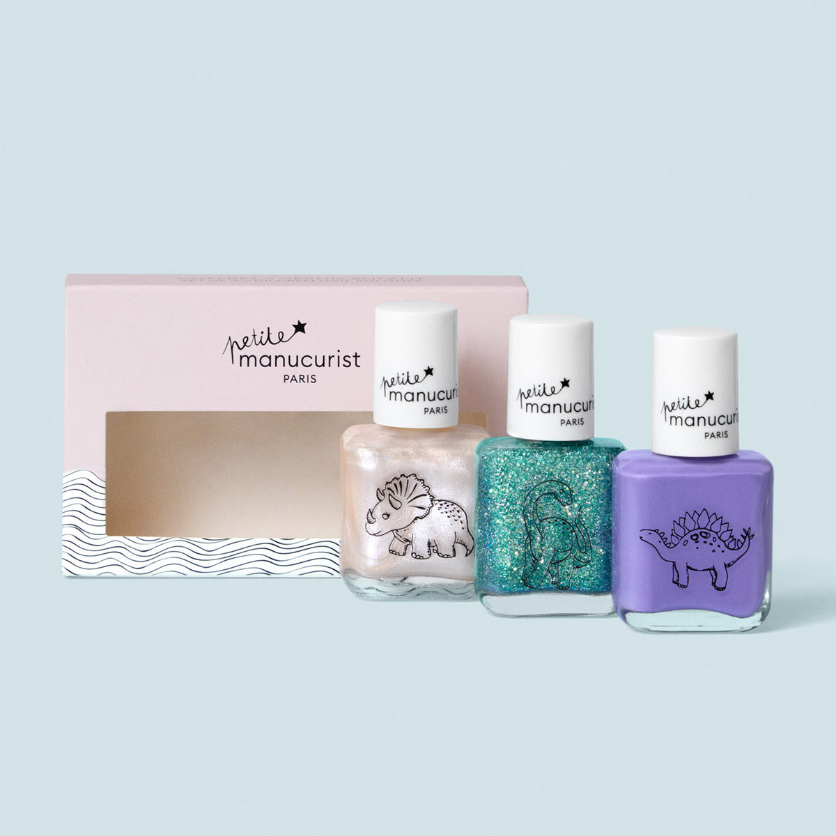 Three bottles from the Manucurist Wild Life Collection—pearl white with triceratops, glittery teal with brontosaurus, and purple with stegosaurus—are displayed before a pink, wavy “petite manucurist Paris” box.