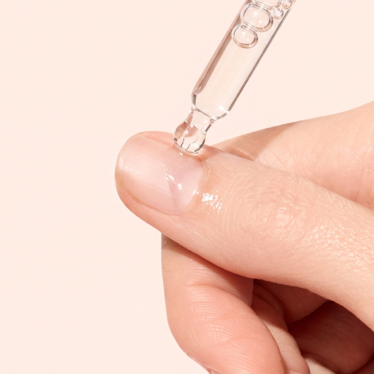 A hand holding a dropper applies a clear liquid to a fingernail against a light beige background.