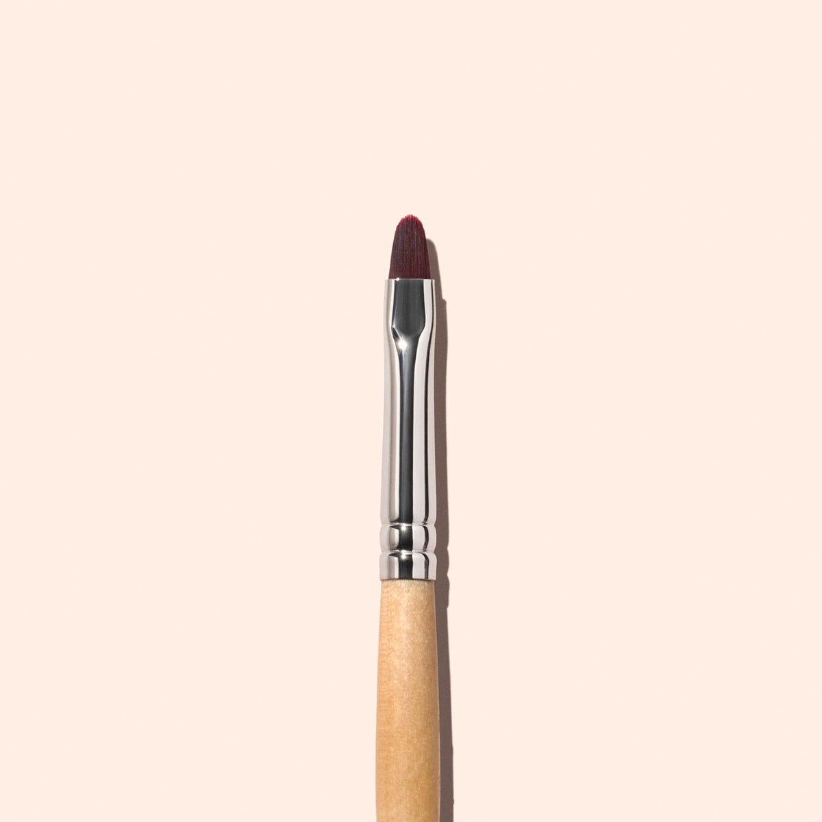 Un pinceau plat Manucurist unique avec un manche en bois et des poils marron est centré verticalement sur un fond uni beige clair, idéal pour une manucure précise et le soin des cuticules.