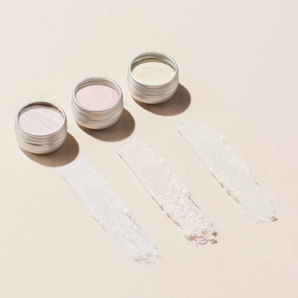 Trois petits pots de Paillettes et Poudres à Effets Manucurist en blanc, rose et crème sont ouverts sur une surface claire, chacun avec un swatch de leur couleur scintillante appliqué devant.