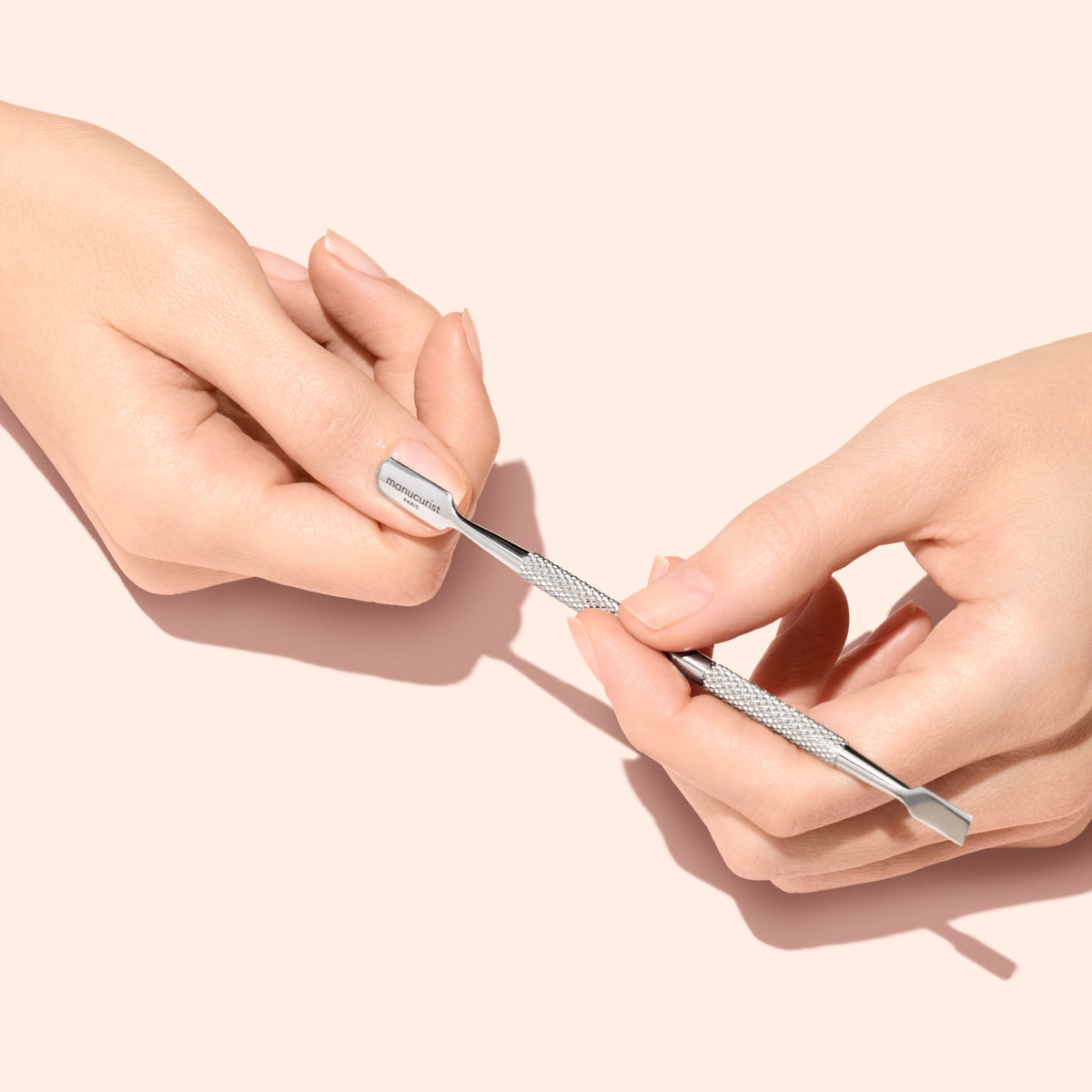 A person uses a metal cuticle pusher to gently push back the cuticle on their fingernail against a light pink background.