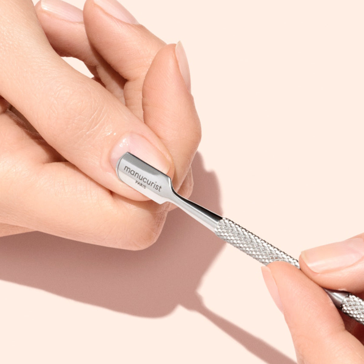 A hand uses a metal cuticle pusher labeled manucurist to gently push back cuticles on another persons fingernail against a light beige background.