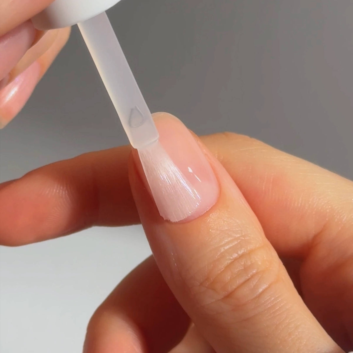 A close-up of a hand applying clear nail polish to a fingernail using a brush. The background is neutral, keeping the focus on the nail and the application process.