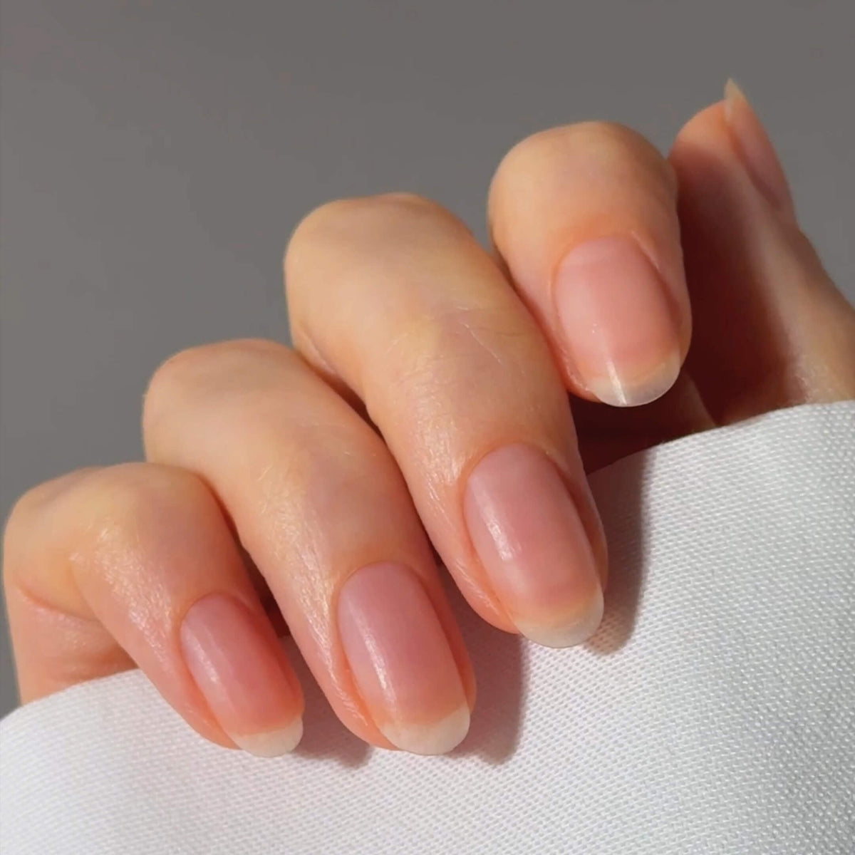 A close-up of a hand with natural, well-groomed fingernails, gently resting on a white fabric. The nails are neatly shaped and have a healthy, glossy appearance.