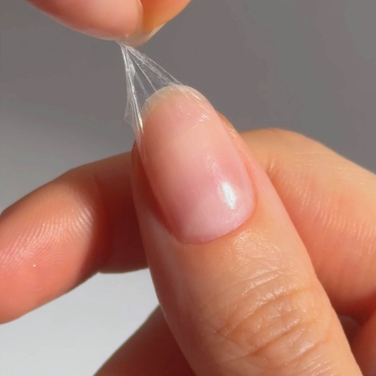 A close-up of a hand peeling a thin, transparent layer off a fingernail, revealing a clean nail underneath.