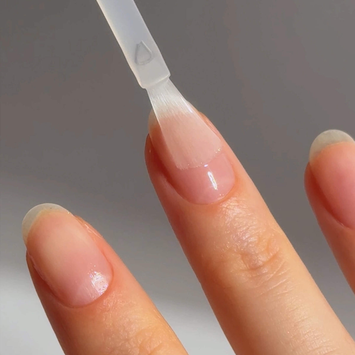 A close-up of a person applying clear nail polish to their fingernail with a small brush. The nails are clean, trimmed, and natural-looking.