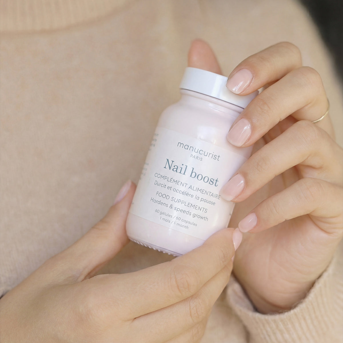 A person with neatly manicured nails holds a bottle of Manucurist Nail Boost food supplements, designed to support nail growth and strength. The background is soft and neutral.