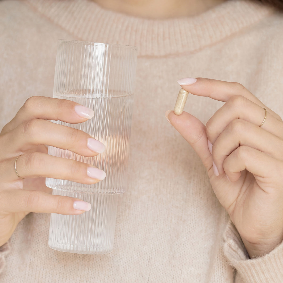 A person in a beige sweater holds a clear glass of water in one hand and a beige capsule pill between their fingers in the other hand.