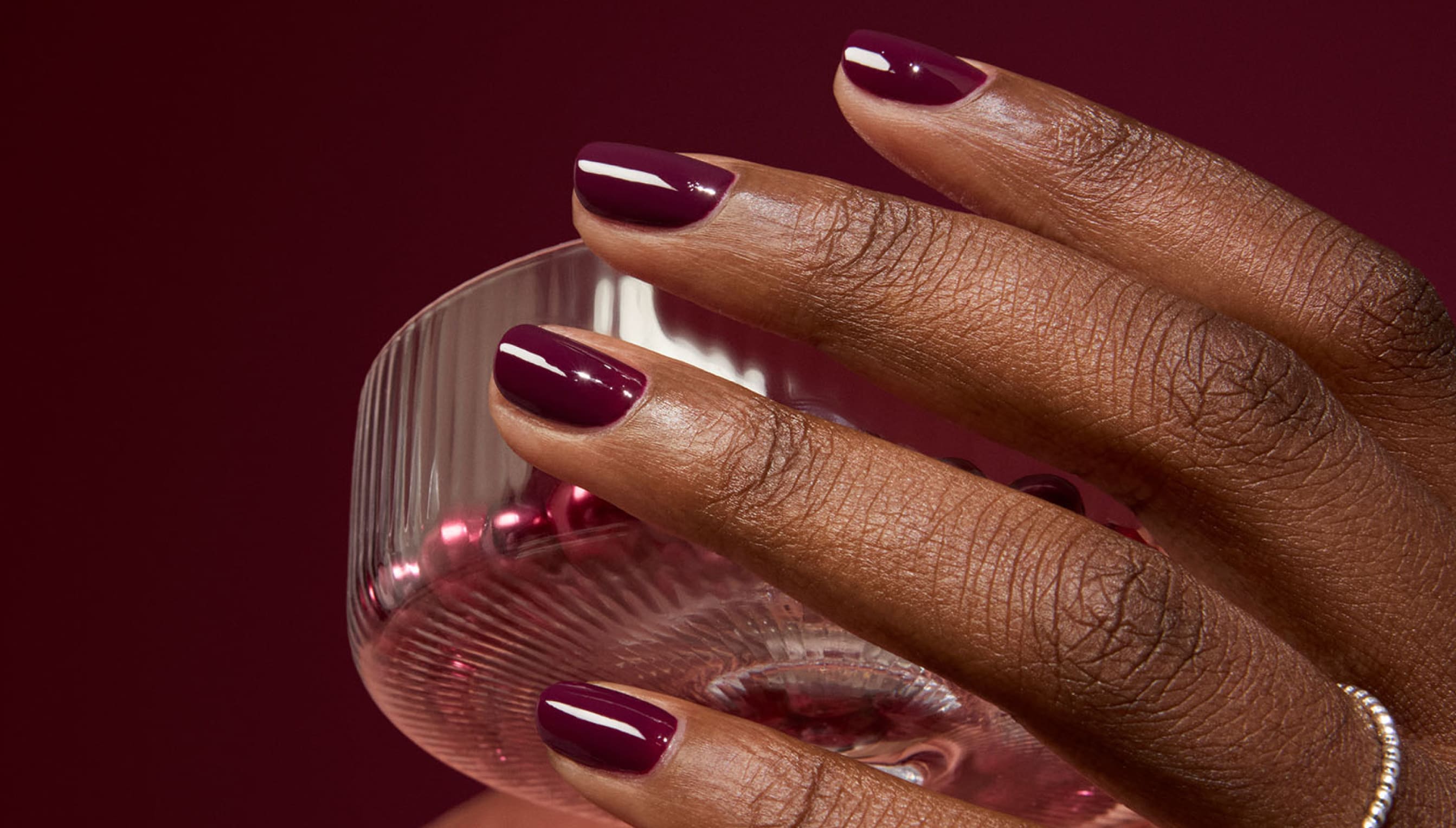 A hand with deep burgundy nail polish holds a textured glass cup against a matching burgundy background. The nails are neatly manicured, and a silver ring is worn on one finger.
