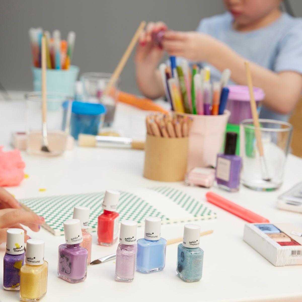 A table with Manucurist’s Joy the Bear Cub kids’ nail polish, pencils, brushes, and art supplies. Two children sit at the table; one in the background plays with craft materials and peach glitter polish made from naturally sourced ingredients.