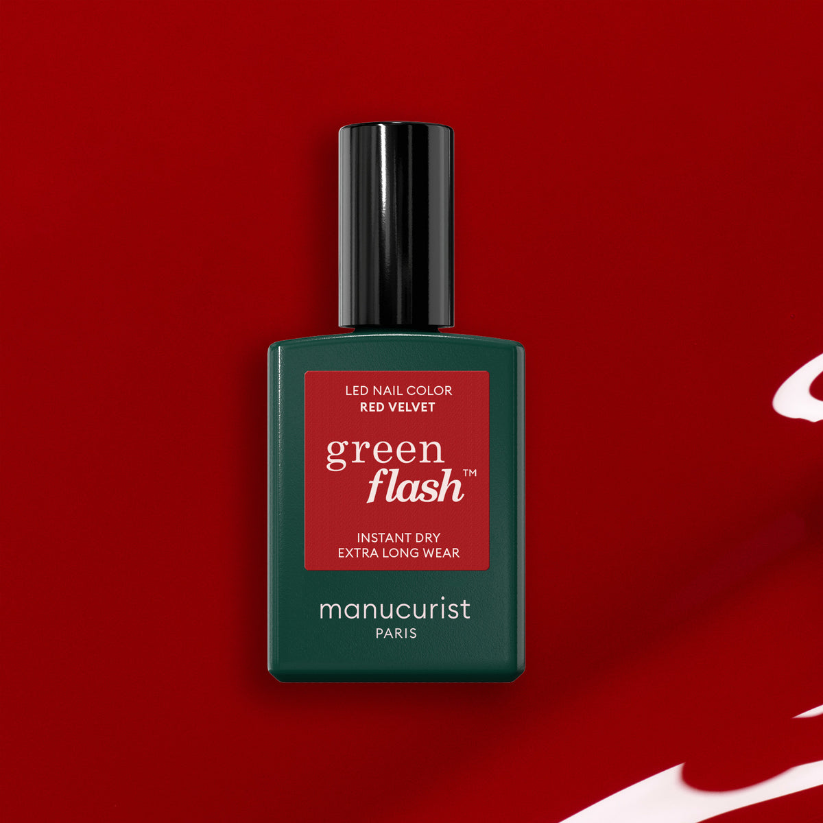 A bottle of Manucurist Red Velvet gel polish sits on a red background, its dark green bottle and red label highlighting the glamorous shade.