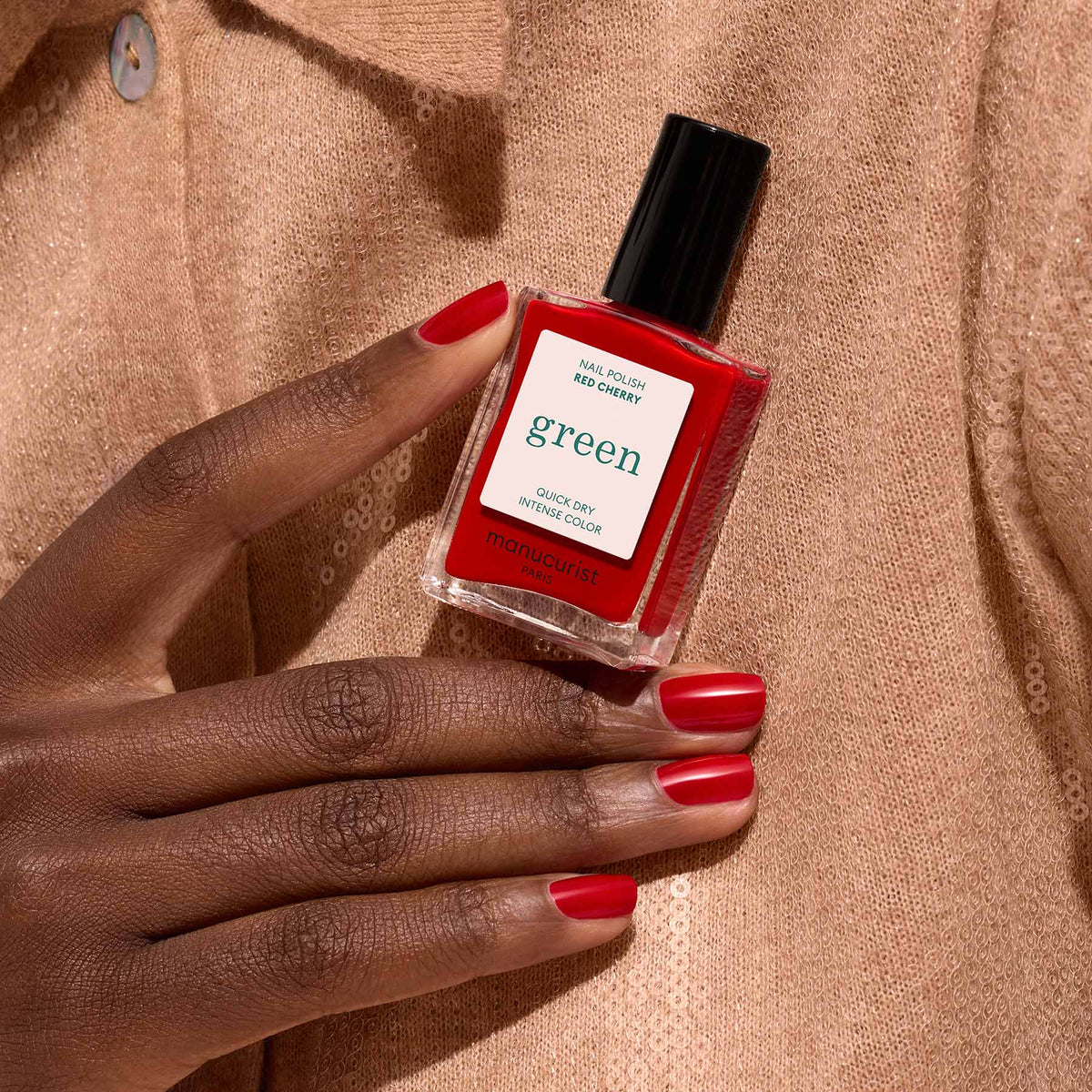 Une main aux ongles rouges soigneusement manucurés tient un flacon de vernis à ongles Manucurist Red Cherry sur un fond en tissu texturé beige.