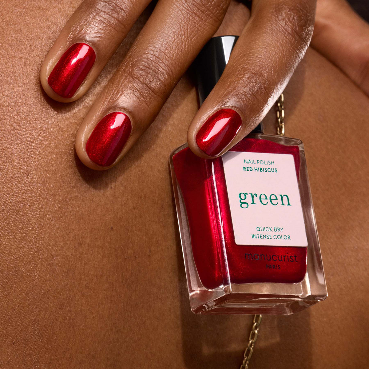 A hand with red-painted nails holds a bottle of Manucurist Red Hibiscus nail polish against brown skin. The vivid, glossy shade makes a bold statement.