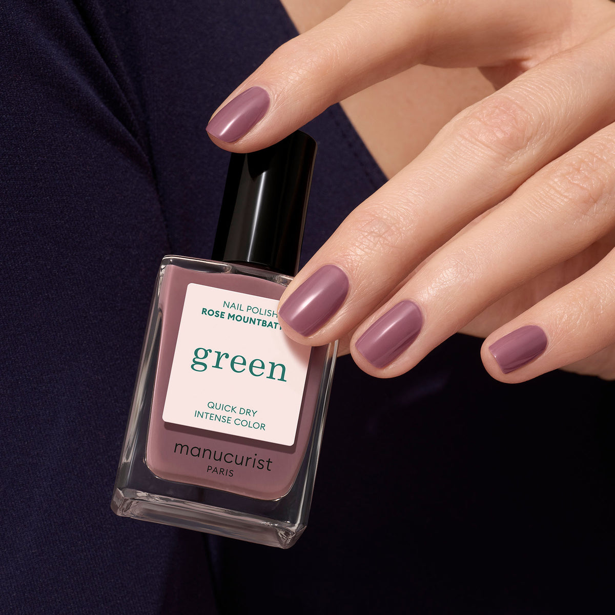 Une main aux ongles courts et soigneusement manucurés, vernis avec la teinte Rose Mountbatten de Manucurist, tient le flacon assorti de la gamme Green™.