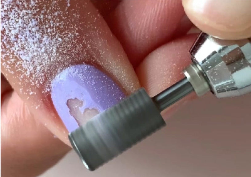 Gros plan sur une ponceuse électrique retirant du vernis à ongles couleur lavande d’un ongle, avec des résidus poudreux visibles sur le doigt.