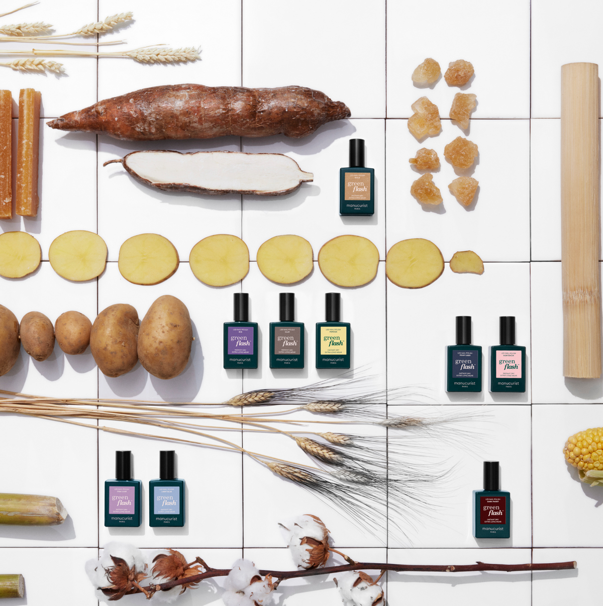 Des flacons de parfum sont disposés en rangées sur une surface carrelée blanche, entourés de pommes de terre, de légumes-racines tranchés, de blé, de maïs, de canne à sucre et de morceaux de sucre, créant un décor naturel et coloré.
