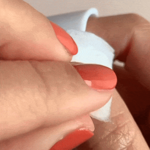A close-up of a hand with coral-colored nails using a white cotton pad to remove nail polish from another fingernail.