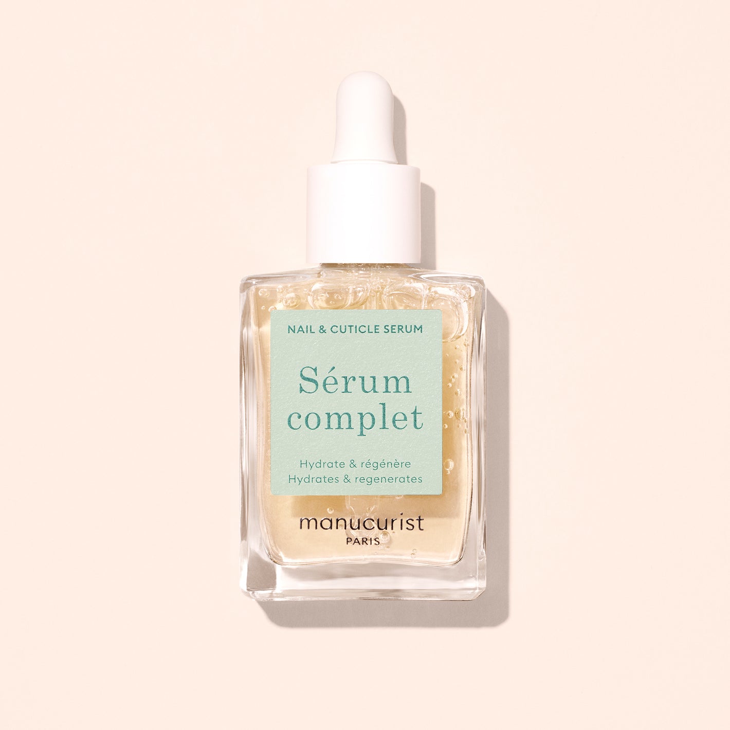 A clear glass dropper bottle labeled Nail Hydrating Serum by Manucurist, containing a nourishing nail serum with pale beige liquid inside, is shown against a light beige background.