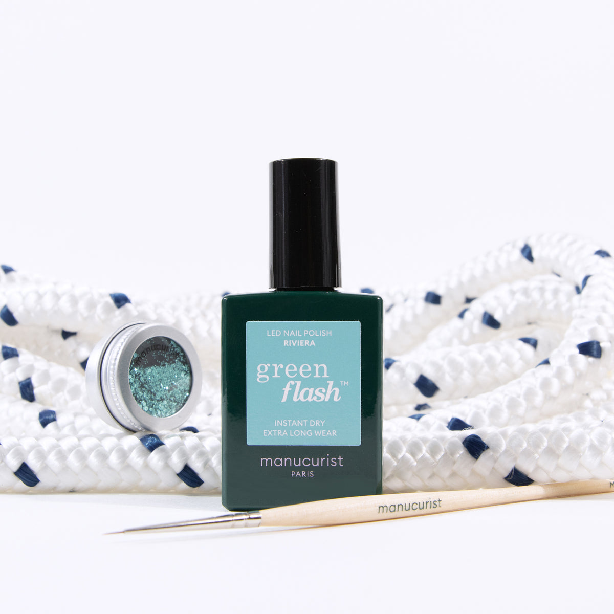 Un flacon de vernis à ongles Underwater Glitter de Manucurist, un petit pot ouvert de paillettes biodégradables et un outil de manucure en bois sont disposés sur une corde blanche enroulée avec des touches de bleu marine.