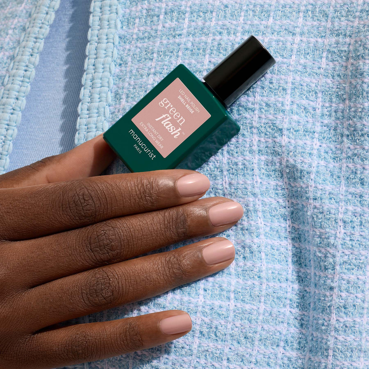 Une main aux ongles Shell Beige soigneusement manucurés tient un flacon de vernis gel Green Flash Manucurist sur un fond de tissu bleu clair texturé.