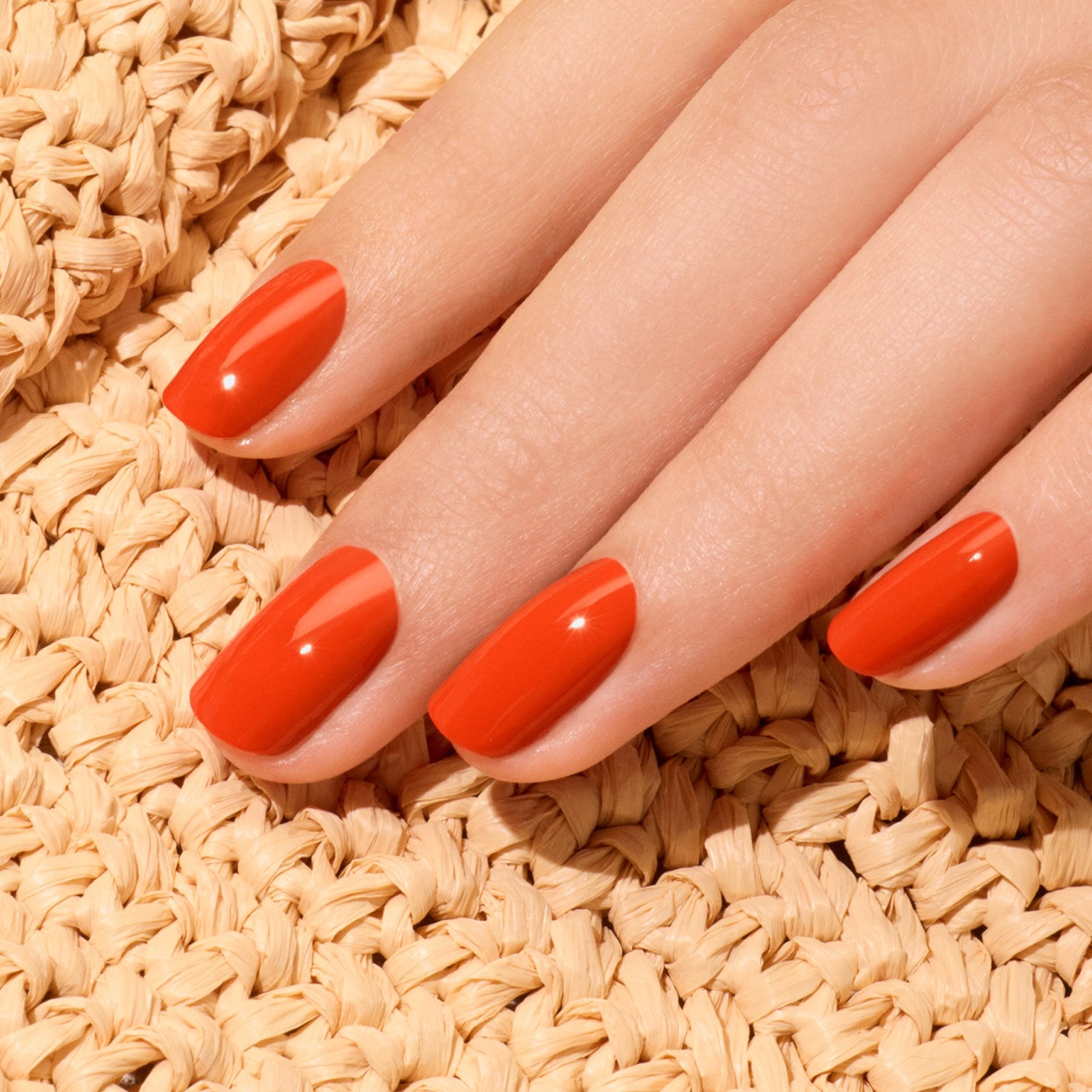 A hand with neatly manicured nails painted in Coral Reef nail polish from Manucurist’s Green™ range rests on a woven beige textured surface.