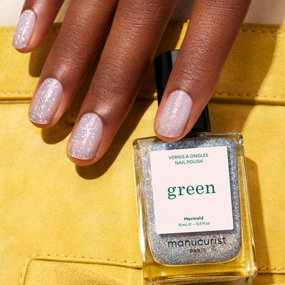 A hand with short, shimmery light pink nails holds a bottle of Manucurist Mermaid nail polish against a yellow background.