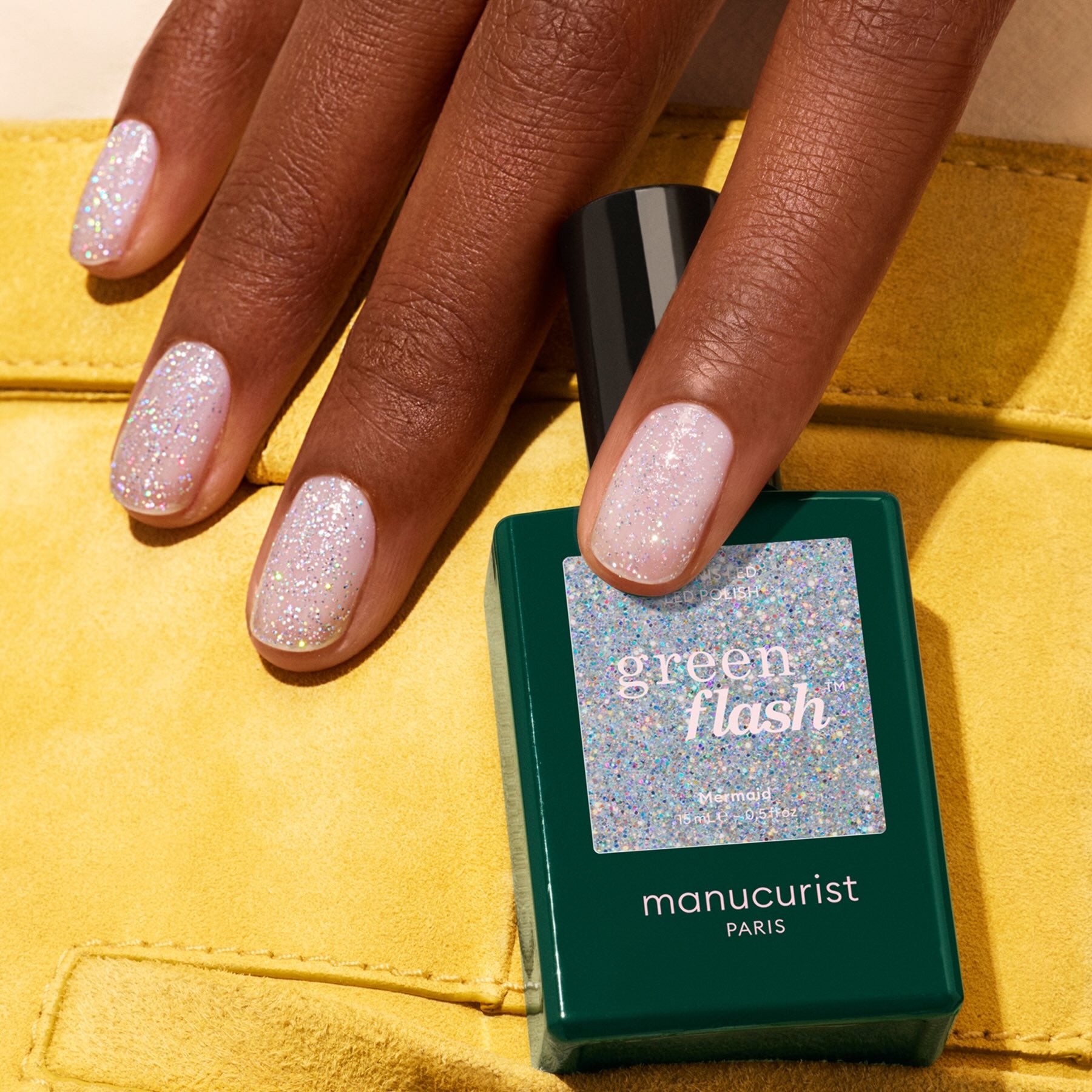 A hand with short, rounded nails painted in sparkly, pastel Mermaid gel polish rests on a yellow surface. A matching bottle of Manucurist Mermaid nail polish is placed nearby.