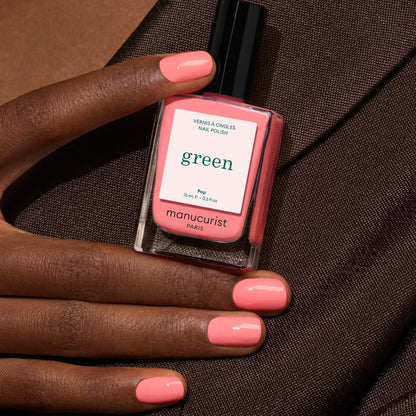 A hand with neatly manicured nails painted in a glossy peach-pink shade holds a matching bottle of Manucurist Pop nail polish against a dark brown fabric background.