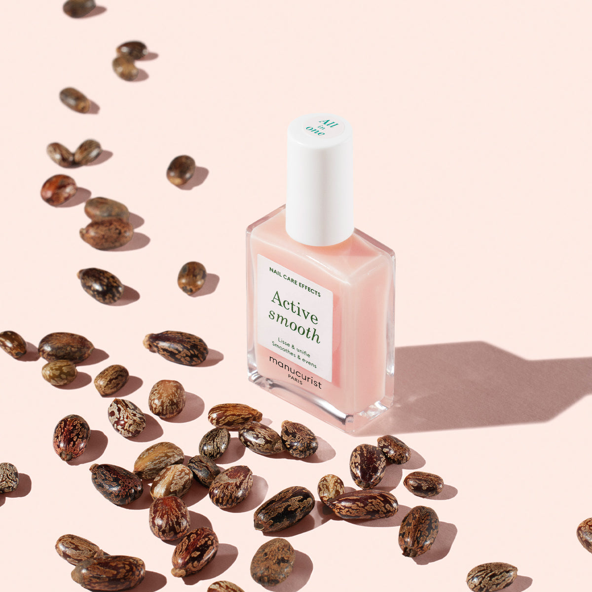 Un flacon de vernis à ongles rose Manucurist Active™ Smooth 00 est posé sur une surface rose clair, entouré de graines brunes éparses et projetant une ombre douce — idéal pour celles et ceux qui recherchent des ongles d&