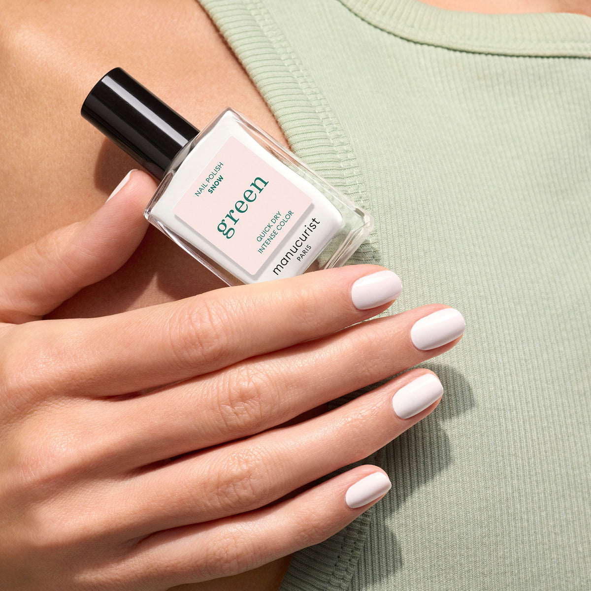 Une main aux ongles soigneusement manucurés rose pâle tient un flacon de vernis à ongles Manucurist Snow de la gamme Green devant un haut côtelé vert sauge. L&