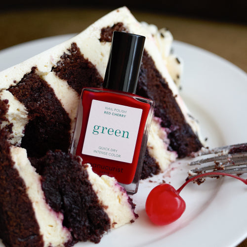 Un flacon de vernis à ongles rouge étiqueté Red Cherry est posé devant une part de gâteau au chocolat avec un glaçage blanc et une cerise au marasquin, avec une fourchette à côté.