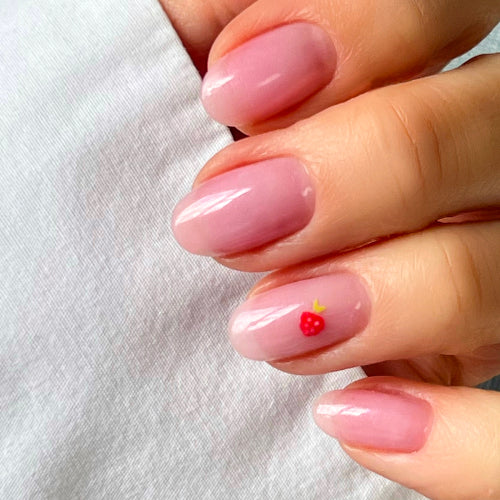Une main aux ongles soigneusement manucurés et vernis d'une teinte rose clair brillante. Un ongle présente un petit motif de fraise rouge avec une tige jaune.