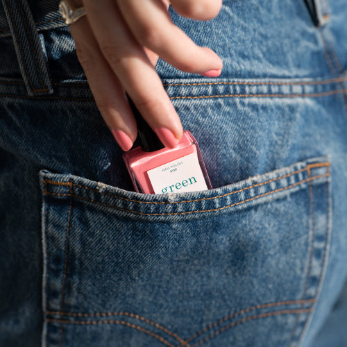 Une main glisse un flacon de vernis à ongles rose étiqueté green dans la poche arrière d'un jean bleu.