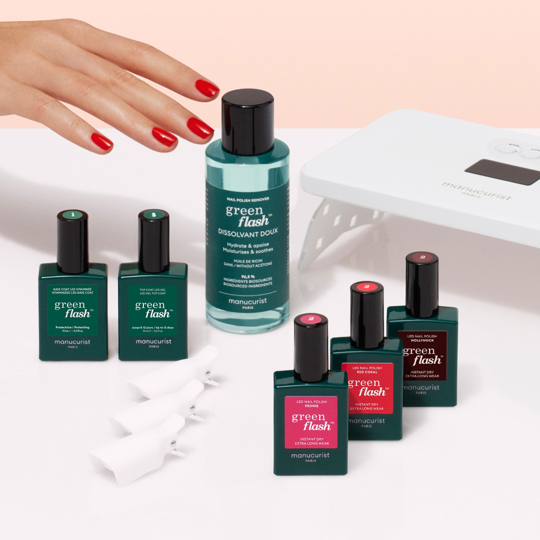 A hand with red-painted nails reaches for the Manucurist Starter Kit, which includes Green Flash polishes, remover, nail clips, and a UV lamp—everything needed for an ultra-glossy manicure—arranged on a white surface.