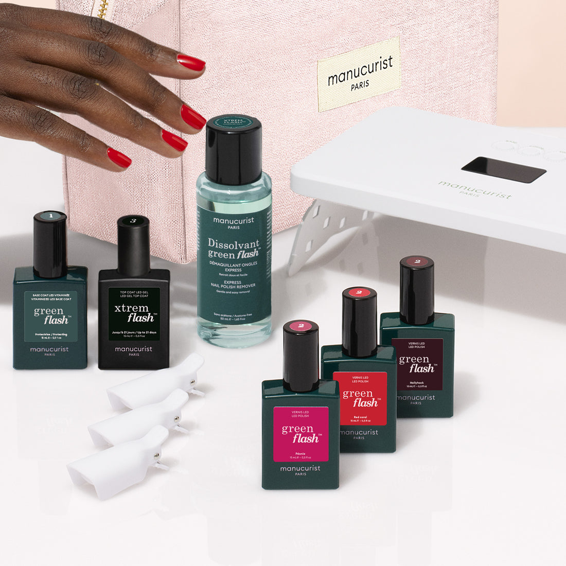 A hand reaches for the Manucurist Starter Kit—featuring a pink gel polish—next to bottles of Manucurist nail polish, a vitaminized base coat, dissolvant, nail clips, and an LED lamp on a white surface.