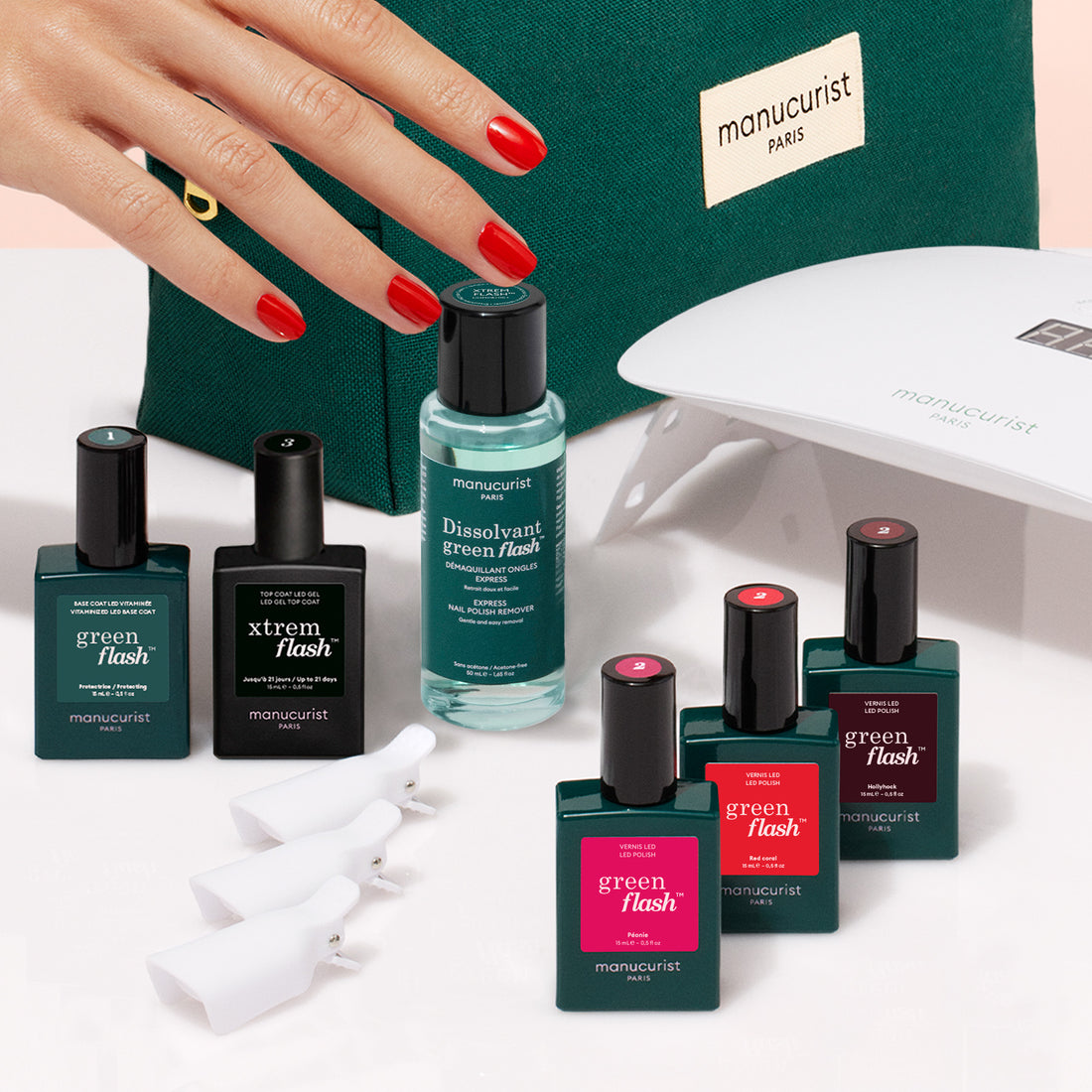 A hand with red polished nails rests on a green Manucurist bag, surrounded by the Manucurist Starter Kit, which includes nail polish, vitaminized base coat, gel polish kit, remover, UV lamp, and care accessories.